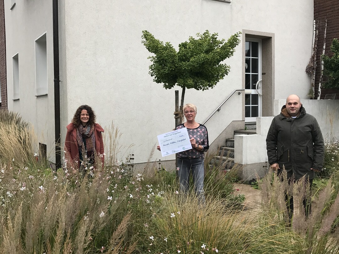 Renate Khte wurde von Marco Weitz (r., VGL NRW) und Miriam Schwenker (l., Landesverband der Gartenbauvereine NRW) in ihrem Vorgarten zur Scheckbergabe besucht.