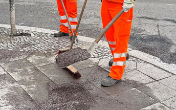 Mitarbeiter reinigen einen Gehweg von Splitt aus dem Winter.